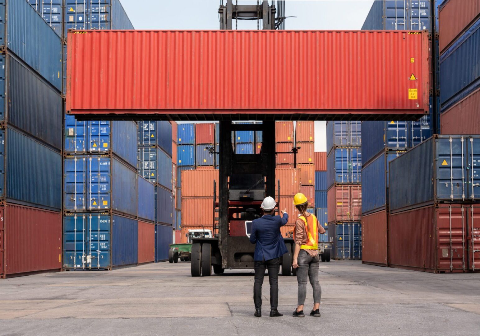 Inspector engineer in hard hat with Female dock worker standing in front of Reach stacker vehicle and used laptop checking cargo freight container to command transportation in logistic shipping yard